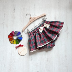 Puede incluir: Un blúmer de bebé de cuadros rojos y verdes con un borde de encaje blanco y un lazo blanco. El blúmer está colgado en una percha de madera con el texto "Colours for Baby" grabado en él. Hay un círculo de madera con el mismo texto y un círculo de vitral de colores.