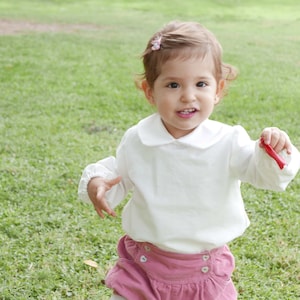 Puede incluir: Una niña pequeña con una camisa blanca de cuello con mangas largas y una falda rosa. La niña está de pie en un campo de hierba y tiene un lazo rosa en el pelo.