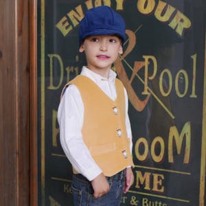 Puede incluir: Un ni&ntilde;o peque&ntilde;o con un chaleco amarillo con botones decorativos, una camisa blanca abotonada, pantalones vaqueros azules y una gorra de newsboy azul. El ni&ntilde;o est&aacute; de pie frente a un letrero que dice "Enjoy Our Drink & Pool Room".