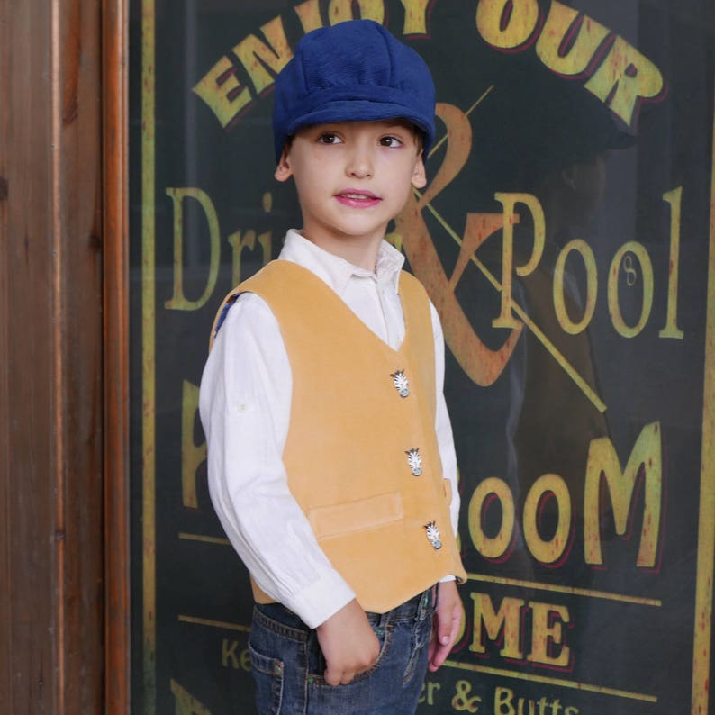 Puede incluir: Un ni&ntilde;o peque&ntilde;o con un chaleco amarillo con botones decorativos, una camisa blanca abotonada, pantalones vaqueros azules y una gorra de newsboy azul. El ni&ntilde;o est&aacute; de pie frente a un letrero que dice "Enjoy Our Drink & Pool Room".