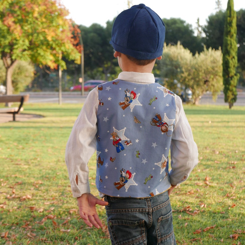 Puede incluir: Una persona joven que lleva un chaleco azul y blanco con estampado de tem&aacute;tica vaquera. El chaleco tiene estrellas, cactus y vaqueros de dibujos animados. La persona tambi&eacute;n lleva un sombrero azul y pantalones vaqueros azules.