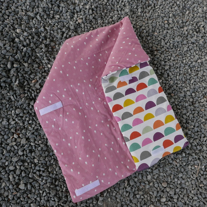 K&ouml;nnte beinhalten: Eine rosa-wei&szlig;e, reversible Babydecke mit einem Tupfenmuster auf der einen Seite und einem bunten geometrischen Muster auf der anderen Seite. Die Decke hat zwei wei&szlig;e B&auml;nder.