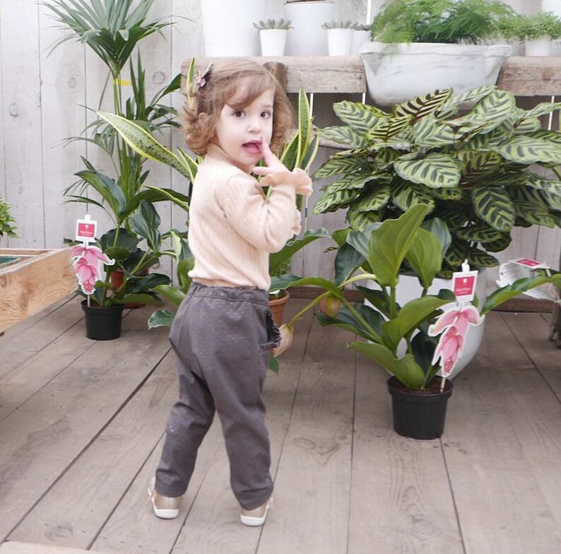 May include: A young girl wearing a light pink sweater and brown pants stands in a greenhouse. She is looking over her shoulder and has her finger in her mouth. There are plants in pots around her.