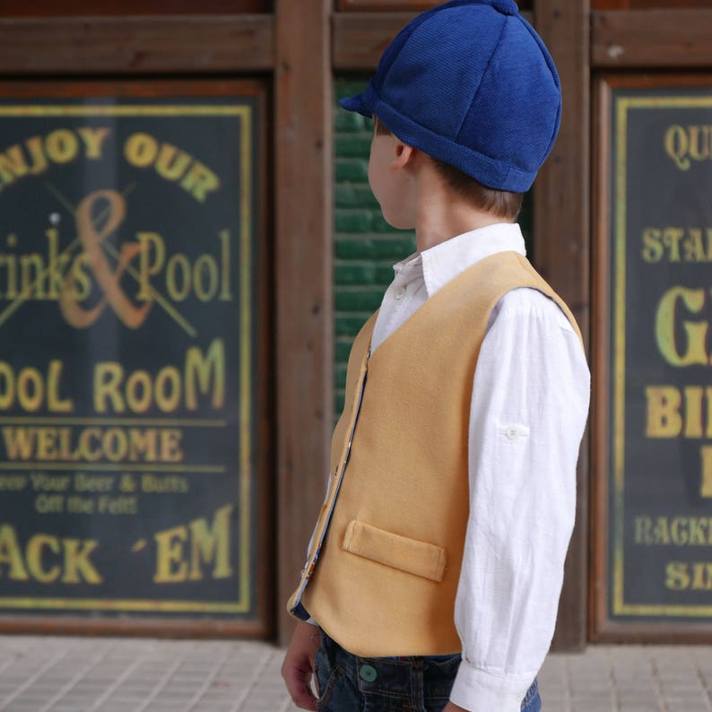 Puede incluir: Un ni&ntilde;o peque&ntilde;o con una gorra de golf azul, una camisa blanca de botones y un chaleco amarillo. Est&aacute; de pie frente a un edificio de madera con un letrero que dice "Enjoy Our Drinks & Pool Pool Room Welcome".