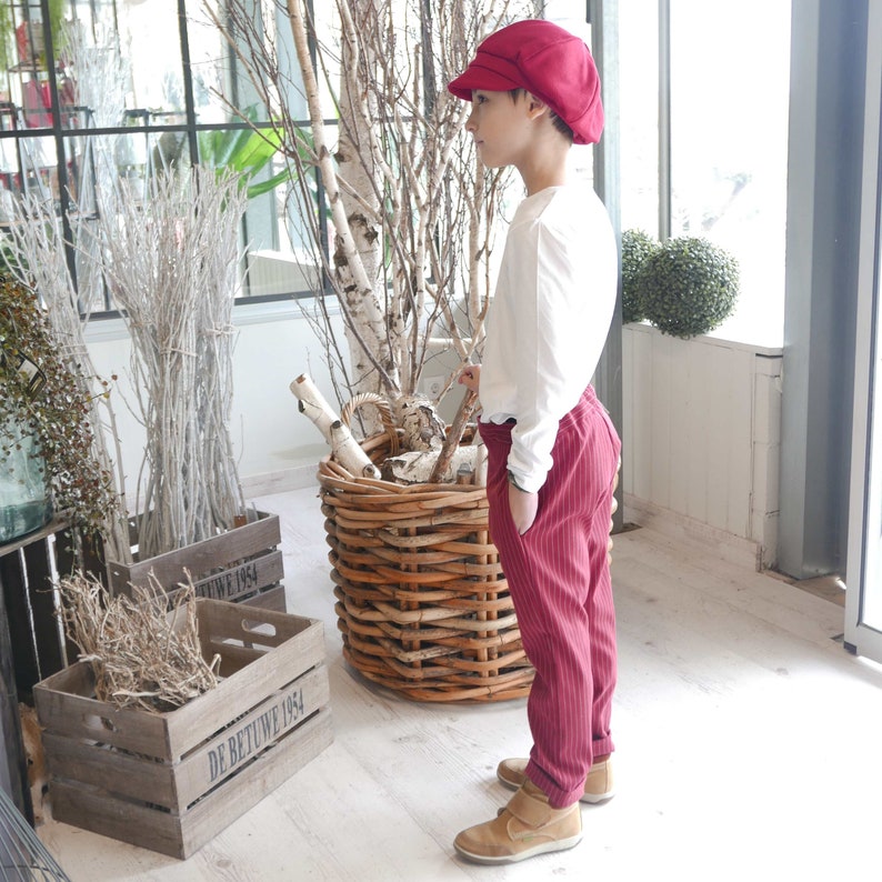 May include: A young person wearing a red and white striped pants, a white long-sleeve shirt, and a red newsboy cap. The person is standing in front of a wooden crate and a wicker basket.
