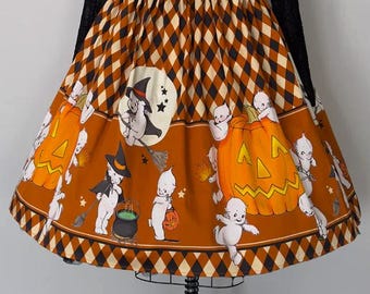 May include: A high-waisted, knee-length skirt featuring a brown and black diamond pattern at the waistband and hem. The main orange panel displays a repeating print of white ghosts, jack-o'-lanterns, witches on broomsticks, and cauldrons.