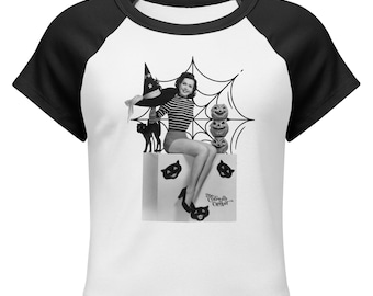 May include: A white raglan t-shirt with black sleeves featuring a black and white graphic of a person in a striped shirt and witch hat sitting on a box with a spiderweb, three stacked jack-o'-lanterns, and the text The Cobweb Casket.