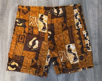 May include: A pair of shorts featuring a patchwork-style print in shades of brown, tan, and cream. The pattern consists of various geometric shapes and repeated graphic illustrations of human footprints, with a single button closure at the waist.