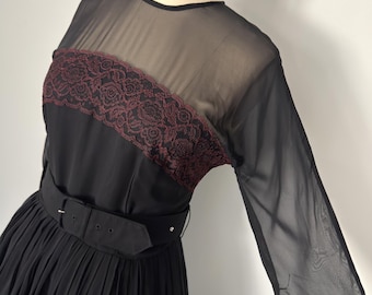 May include: A black dress featuring sheer long sleeves and a sheer upper bodice panel. A dark red floral lace trim accents the chest area. The dress includes a wide black fabric belt with a rectangular buckle and a pleated skirt section.