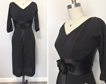 May include: A black knee-length dress featuring a V-neckline, three-quarter length sleeves, and a wide satin waistband adorned with a fabric rose. The dress includes a vertical satin panel extending from the waist and the text The Cobweb Casket.
