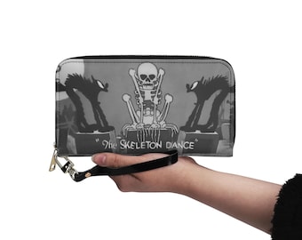 May include: A rectangular black and white wallet featuring a graphic print of a skeleton sitting between two arched-back cats. The text "The SKELETON DANCE" is printed at the bottom. The wallet includes a black wrist strap attached to a metal zipper.