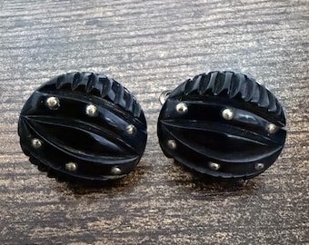 May include: A pair of oval-shaped black earrings featuring a carved, ribbed texture and small, embedded silver-toned metal studs arranged in a symmetrical pattern across the surface of each piece, showcasing a dark, polished finish.