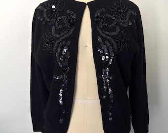 May include: A black long-sleeve cardigan featuring intricate black beadwork and sequin embellishments in a floral-inspired pattern along the front panels. The garment has a round neckline and is displayed on a dress form with the text COLLAPSIBLE SHOULDER 6.