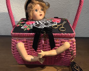 May include: A pink woven basket with a handle, featuring a doll head emerging from the top and two doll arms protruding from the front. The lid has a black fabric cover with pink roses, a black bow, and small scissors. A tag reads Doll Parts.