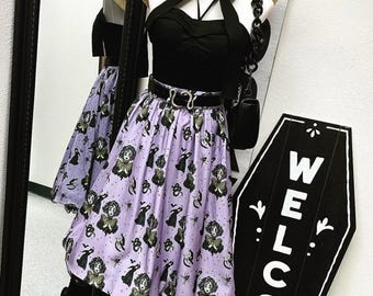 May include: A mannequin wearing a black halter top, a wide-brimmed straw hat, and a lavender skirt featuring a repeating print of figures, cats, and spiders. A black belt with a silver snake buckle and a black handbag are included.