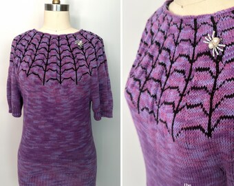 May include: A short-sleeved knit sweater in variegated shades of purple and pink, featuring a black spiderweb pattern around the neckline and a silver-toned spider brooch with a pearl-like center and clear rhinestones pinned to the upper chest.