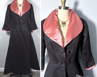 May include: A long black coat featuring a wide, satin-finish pink shawl collar and matching pink cuffs. The coat has three-quarter length sleeves, a fitted waist, and four black fabric-covered buttons. A gold-toned necklace is visible at the neck.