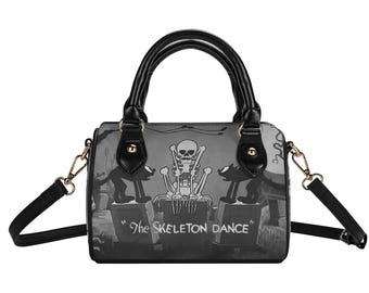 May include: A black and white handbag featuring a graphic print of a skeleton sitting on a tombstone between two black cats. The text "The SKELETON DANCE" is printed on the front. The bag has black handles, gold-tone hardware, and a detachable strap.