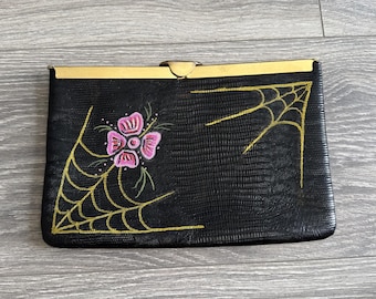 May include: A black clutch bag with a textured lizard-skin pattern, featuring a gold-tone metal frame closure. The front is decorated with hand-painted gold spider webs in the corners and a central pink flower with green accents on the left side.