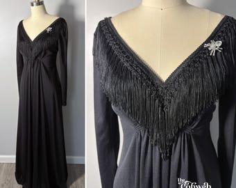 May include: A floor-length black dress with long sleeves and a V-neckline. The bodice features black fringe trim and a silver-toned spider brooch. The fabric is solid black with a flowing skirt. The text The Cobweb Casket is visible at the bottom.