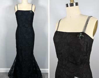 May include: A floor-length black lace dress featuring a square neckline, thin rhinestone-encrusted straps, and a flared mermaid-style hem. A small green spider-shaped brooch is pinned to the upper left chest area of the bodice.