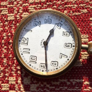 May include: Antique pocket watch with a white face, black numerals, and black hands. The watch has a gold-colored metal casing and a winding stem. The face reads "3 Days". The watch is set against a red and white patterned background.