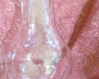 May include: A clear glass lamp shade with a rounded base and a flared top. The shade has a smooth, curved design and is likely for a vintage or antique lamp. The background is a patterned pink fabric.
