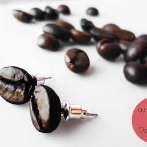 Könnte beinhalten: Ohrstecker in Form von Kaffeebohnen mit silbernen Stiften, präsentiert auf einer weißen Oberfläche mit verstreuten Kaffeebohnen. Die Ohrringe sind dunkelbraun und haben eine strukturierte Oberfläche. Ein roter Kreis mit weißem Text befindet sich in der unteren rechten Ecke.