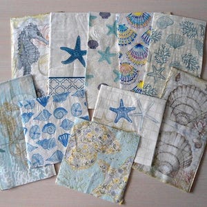 May include: A collection of decorative paper napkins with various sea-themed designs. The napkins feature images of seahorses, starfish, seashells, and coral in shades of blue, white, and yellow. The napkins are arranged on a light-colored surface.