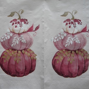 May include: Two decorative napkins featuring a stacked pumpkin design. The pumpkins are in shades of burgundy, pink, and white, with green and gold leaf accents. The napkins are on a light pink background.
