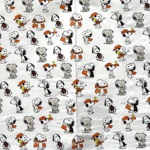 May include: A white paper napkin featuring a repeating pattern of Snoopy and Woodstock in various Halloween costumes, including a mummy, a vampire with an orange cape, and a pirate with an orange bandana, all rendered in black, white, and orange.