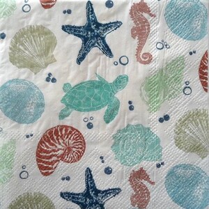 May include: White napkin with a repeating pattern of sea creatures. The design includes a teal sea turtle, navy blue starfish, coral seahorses, and various seashells in teal, coral, and green. Bubbles are scattered throughout the design.