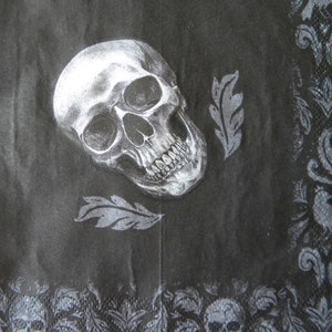May include: A black paper napkin featuring a detailed white illustration of a human skull positioned centrally. Two stylized gray leaves are placed near the skull. The border displays a repeating pattern of dark gray floral motifs and skulls.