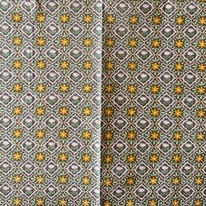 May include: A folded paper napkin with a repeating floral pattern. The design features yellow flowers, white blossoms, and green leaves set against a dark gray background. The pattern is arranged in a diamond grid.