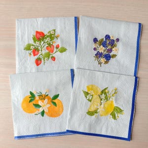 May include: Four white paper napkins with colorful fruit and floral designs. Each napkin features a different design: strawberries, purple flowers, oranges, and lemons. The napkins have a blue border.