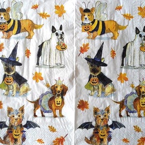 May include: White paper napkins with various dog illustrations dressed in Halloween costumes. The dogs are dressed as a bee, ghost, witch, princess, and bat. Each dog is holding a pumpkin-shaped treat bucket. Autumn leaves are scattered around the dogs.