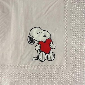 May include: A white paper napkin featuring a Snoopy cartoon character holding a red heart. The napkin has a decorative border with a diamond pattern. The Snoopy image is in black, white, and red.