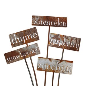 May include: A collection of weathered metal garden markers with the names of various plants in white lettering. The markers include "thyme", "strawberries", "watermelon", "raspberry", and "zucchini". The metal has a rusty, aged appearance.