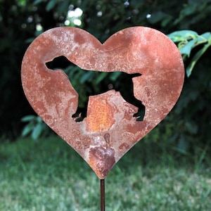 May include: A heart-shaped, rusty metal garden stake featuring a silhouette of a dog. The dog is standing on a small platform within the heart. The stake is in a grassy area with a blurred green background.