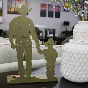 May include: A decorative metal sculpture depicting a father and child holding hands, both wearing cowboy hats. The sculpture is a muted gold color and stands on a rectangular base. Two white vases with diamond patterns are also visible.
