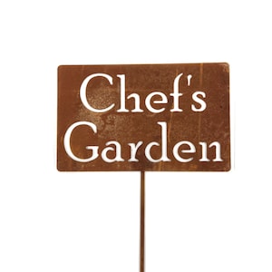 May include: A rusty brown metal sign with the words "Chef's Garden" in white lettering.
