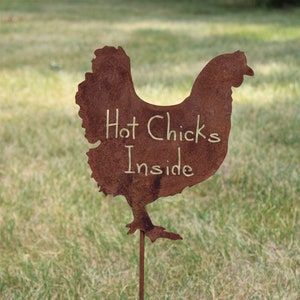 May include: A rusty metal chicken silhouette with the words "Hot Chicks Inside" written on it. The chicken is on a metal stake and is standing in a grassy field.