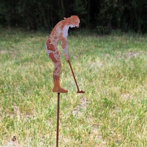May include: A rusty metal golfer silhouette, with a golf club, is staked into the ground. The figure is in a putting stance, with a weathered, textured appearance. The sculpture is set in a grassy field.