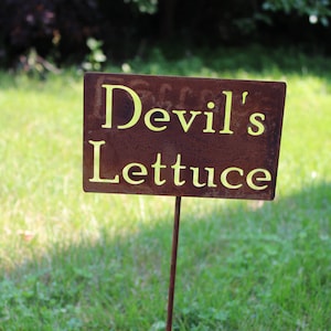 May include: A rusty metal sign with yellow lettering reads "Devil's Lettuce" and is stuck into the ground in a grassy area.
