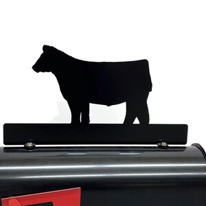 May include: Black metal mailbox with a black cow silhouette. The mailbox has a red mail slot. The cow silhouette is mounted on a black rectangular bar. The mailbox is black.