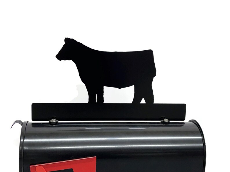 May include: Black metal mailbox with a black cow silhouette. The mailbox has a red mail slot. The cow silhouette is mounted on a black rectangular bar. The mailbox is black.