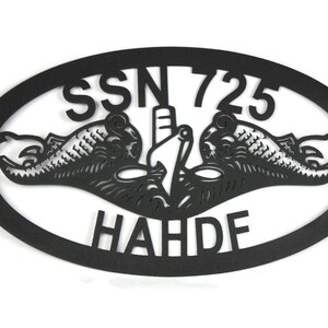 Puede incluir: Un letrero de pared ovalado de metal negro con un dise&ntilde;o recortado de la insignia de guerra submarina, que incluye dos delfines flanqueando la proa de un submarino. El texto SSN 725 est&aacute; sobre la insignia y el texto HAHDF est&aacute; debajo.