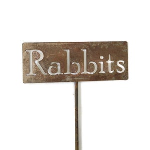 May include: A rectangular, weathered metal garden stake with the word "Rabbits" cut out in a serif font. The stake is brown with a rusty appearance and is designed to be placed in the ground.