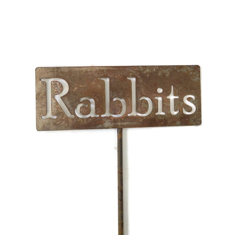 May include: A rectangular, weathered metal garden stake with the word "Rabbits" cut out in a serif font. The stake is brown with a rusty appearance and is designed to be placed in the ground.