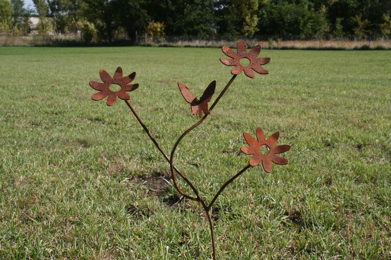 以下が含まれることがあります： 3つの花の咲く花と蝶をあしらった、さびた金属製の庭の彫刻。花の中心には丸い切り抜きがあります。彫刻はさびた金属でできており、草地の中に設置されています。